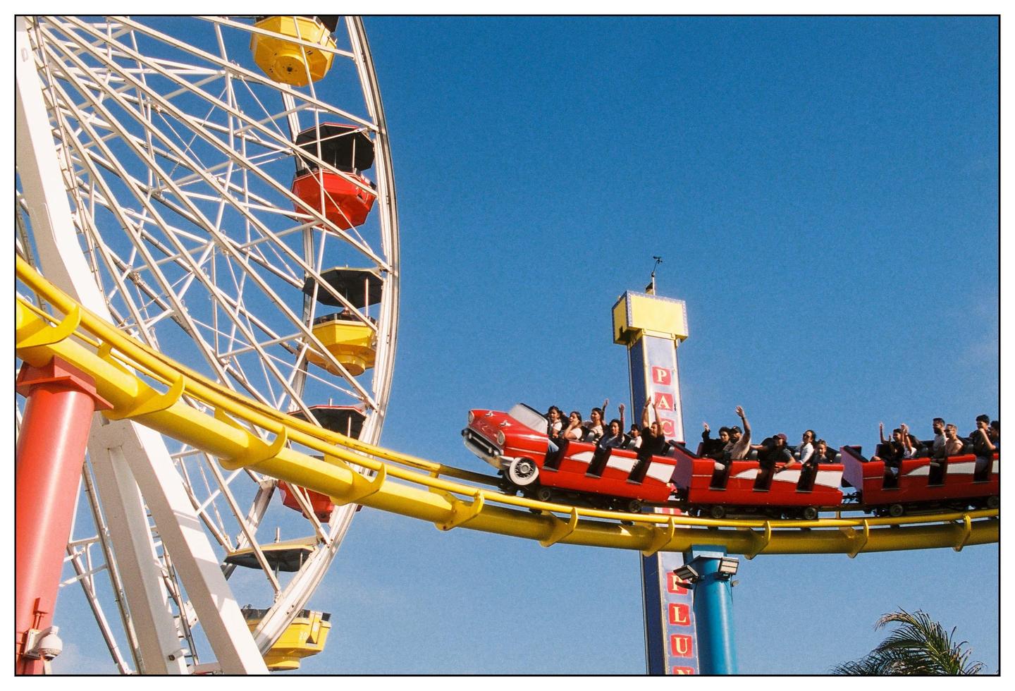 A red roller coaster car full of people with their hands in the air passes a ferris wheel against a clear blue sky.