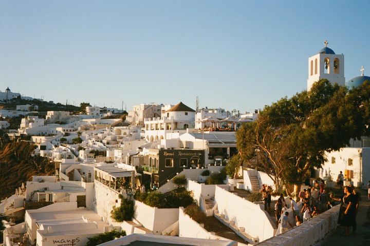 A hillside town with white buildings and a blue-domed church is bustling with people on a sunny day.