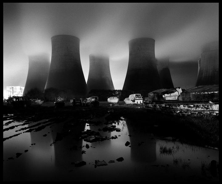 Several cooling towers emit steam into a hazy sky above a muddy, reflective foreground with abandoned boats.