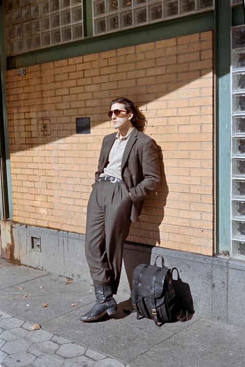 A man wearing sunglasses, a suit jacket, and cowboy boots leans against a brick wall with a backpack on the ground beside him.