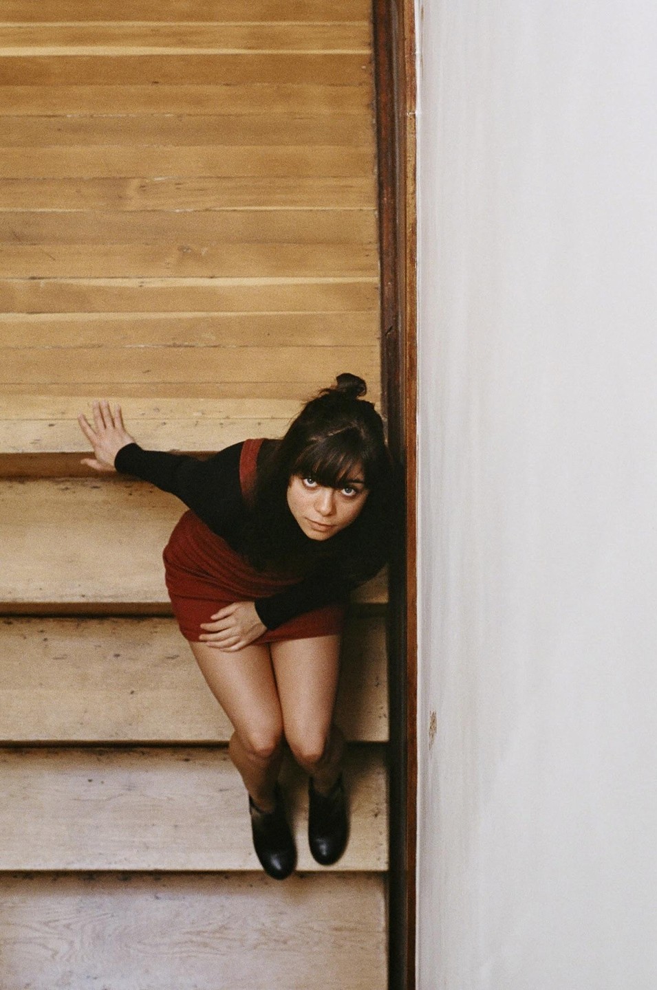 A woman with bangs and a bun looks up at the camera while sitting on wooden stairs.