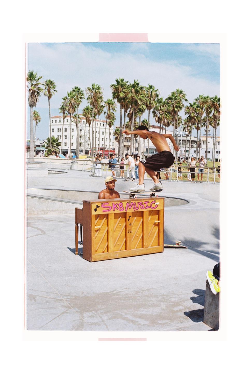 A man on a skateboard jumps over a piano in a skate park with palm trees and buildings in the background.