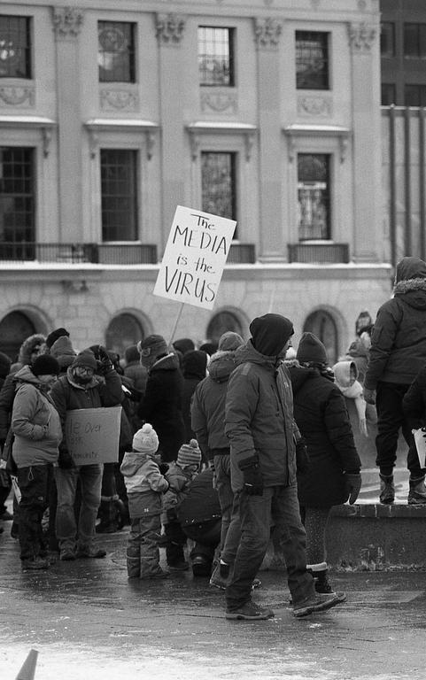 A crowd of people, including children, are gathered on a street with a building in the background, and one person holds a sign that reads "The MEDIA is the VIRUS."