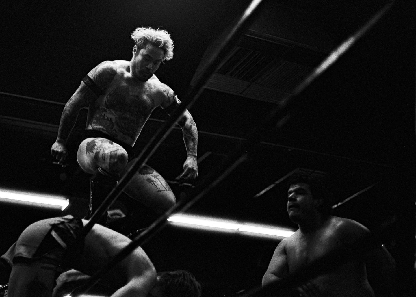 A tattooed wrestler stands on the top rope of a wrestling ring, looking down at another wrestler on the mat, with a third wrestler in the background.