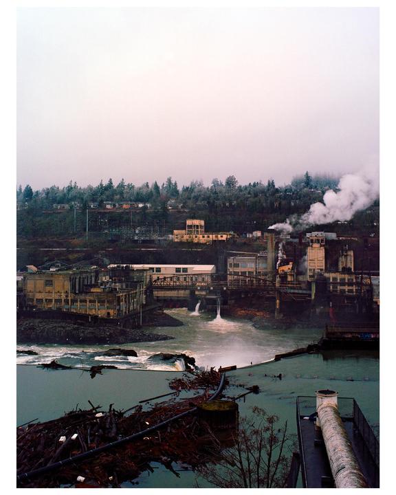 A river flows past industrial buildings and a forested hill under a hazy sky.