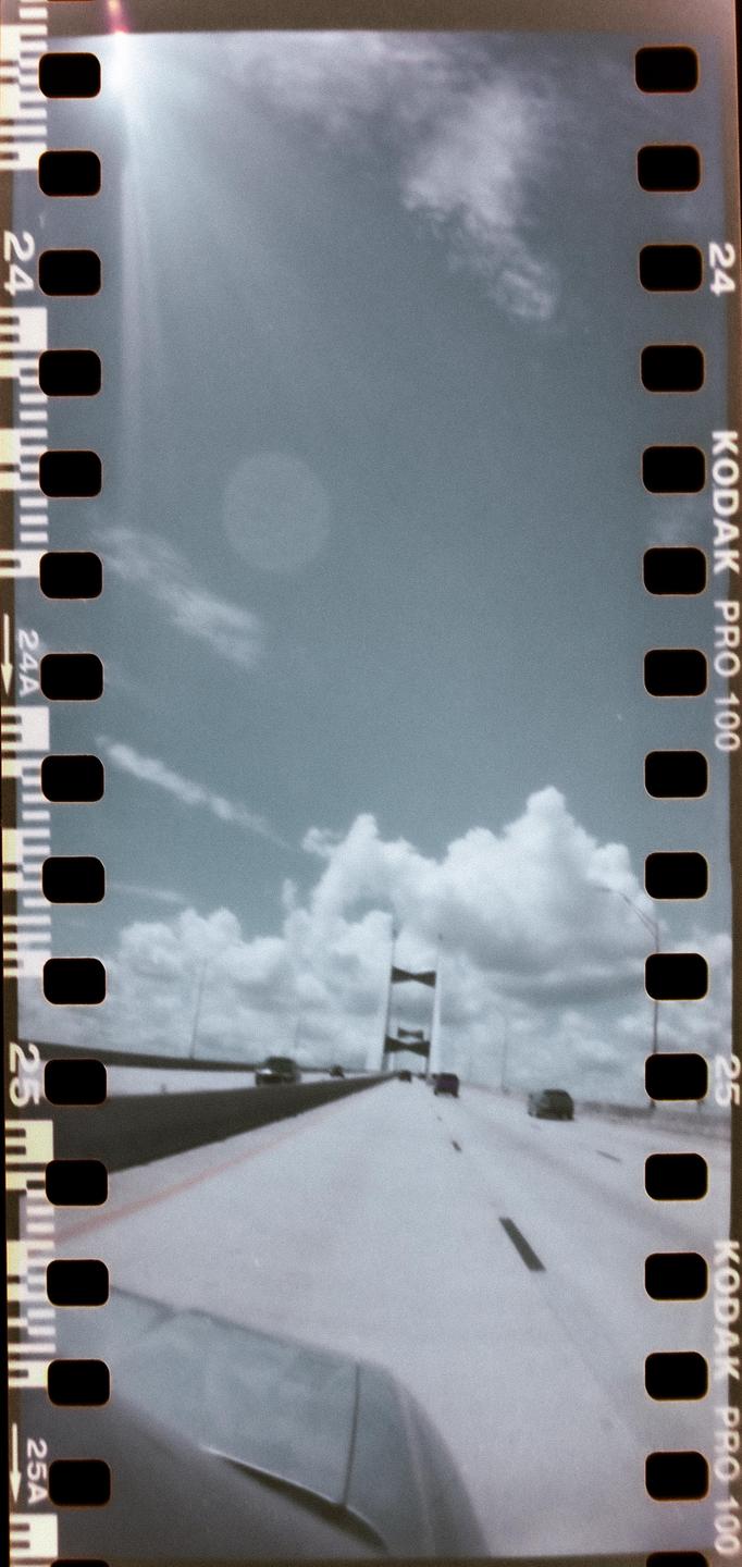 A bridge is visible in the distance from a car driving on a highway under a cloudy sky.