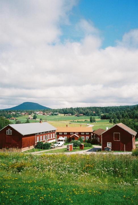 Red farm buildings are nestled in a green valley with a mountain in the distance under a cloudy sky.