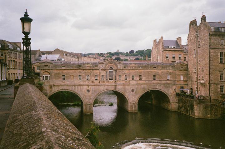 A stone bridge with multiple arches spans a river, surrounded by historic buildings under a cloudy sky.
