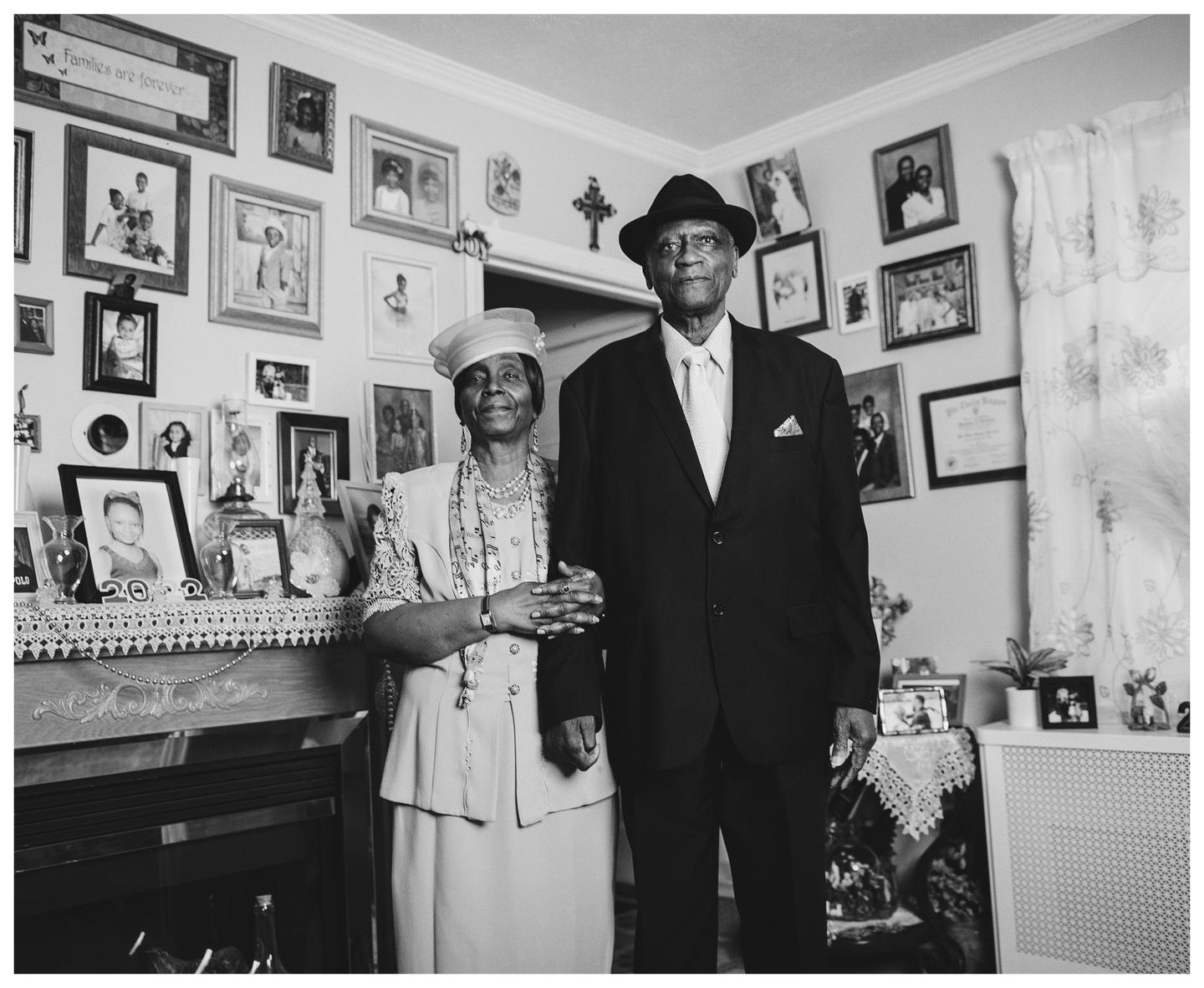 A man and a woman stand arm in arm in a room filled with framed photographs.