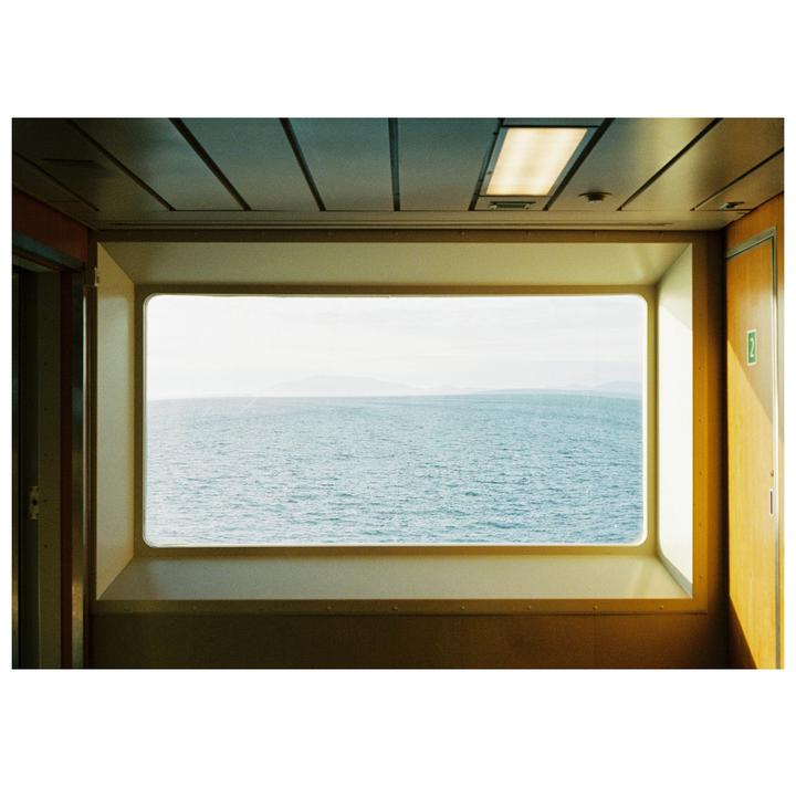 A rectangular window on a boat looks out onto a vast expanse of ocean with distant land visible under a bright sky.