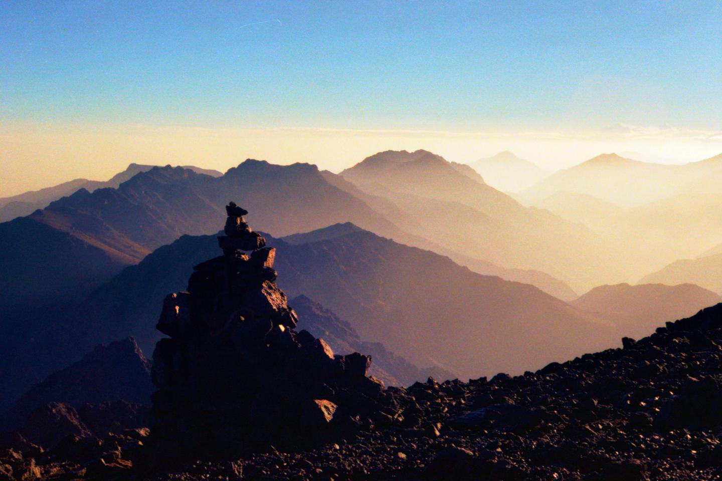 A cairn stands on a rocky mountain peak with many layers of mountains in the background under a hazy sky.