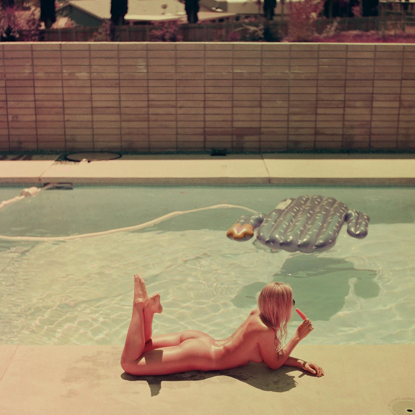 A woman lies nude on the edge of a swimming pool, eating a popsicle, with an inflatable cactus floating in the water.