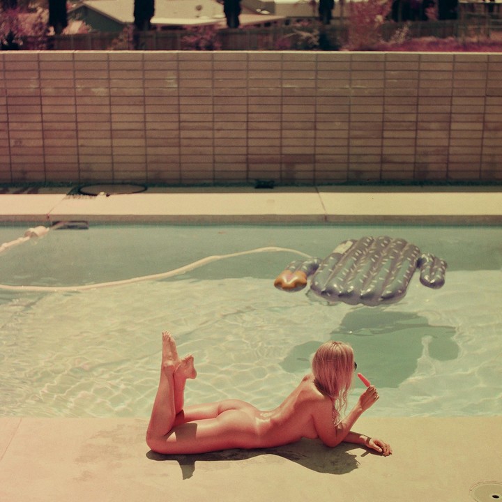 A woman lies nude on the edge of a swimming pool, eating a popsicle, with an inflatable cactus floating in the water.