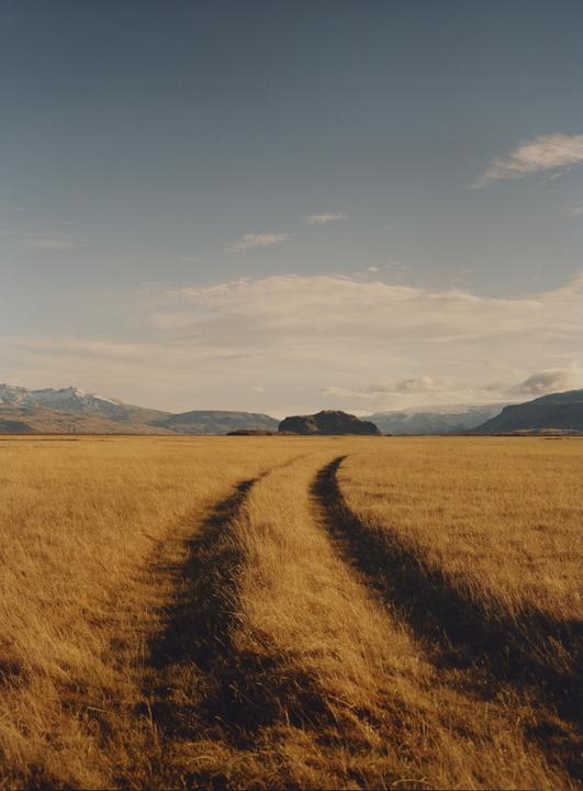 A dirt track curves through a field of golden grass towards mountains under a partly cloudy sky.