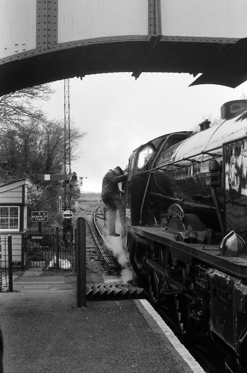 A man climbs into the cab of a steam train at a station next to a signal box.