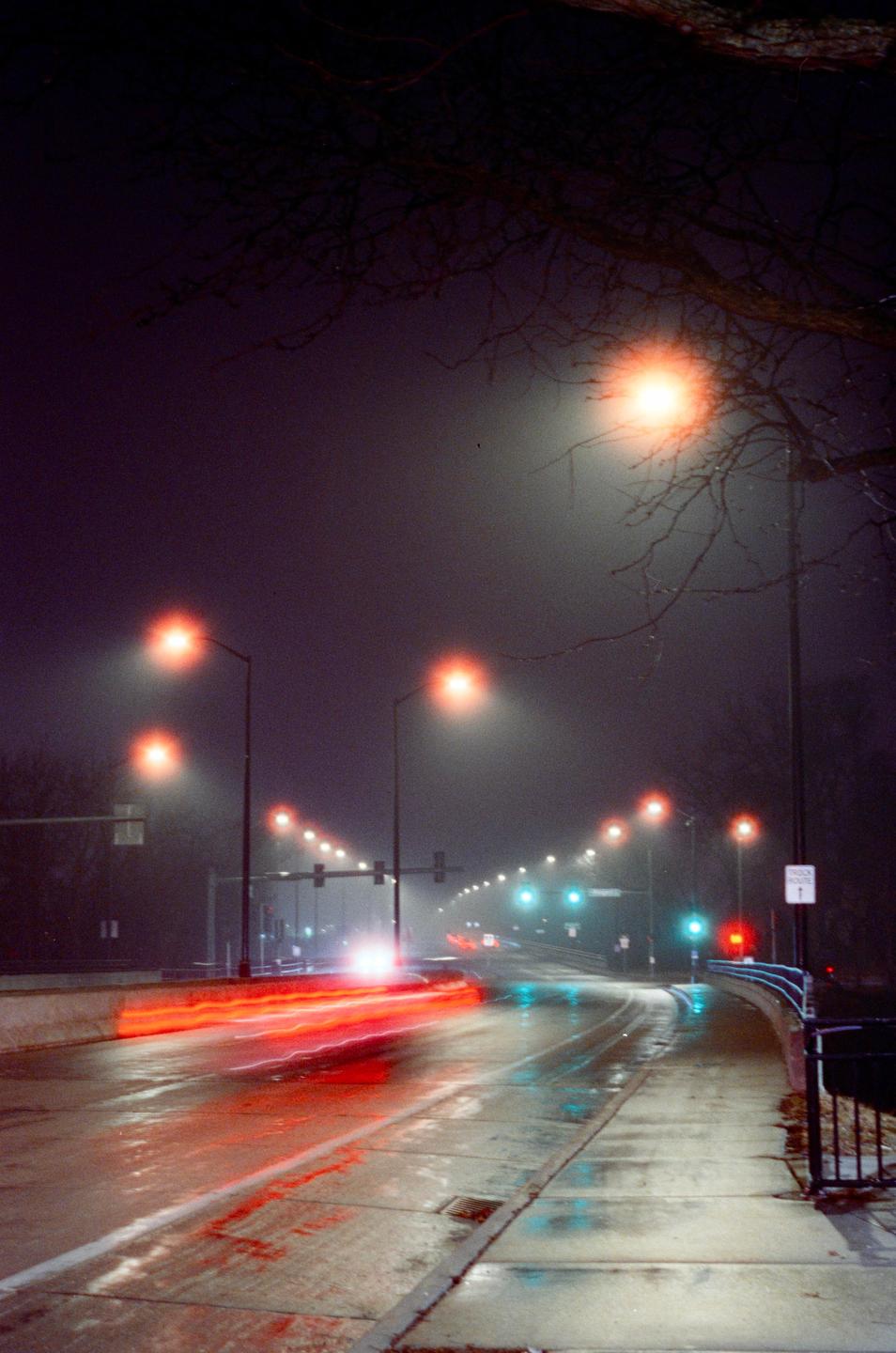 A wet road at night is illuminated by streetlights and the light trails of passing vehicles.