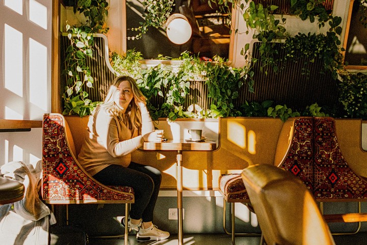 A woman sits at a table in a cafe with plants on the wall and sunlight streaming through a window.
