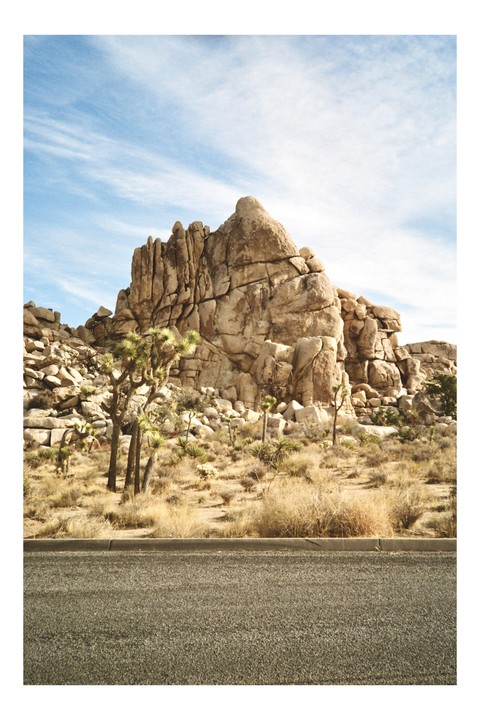 A large rock formation stands behind a desert landscape with Joshua trees and dry brush, with a road in the foreground under a blue sky.