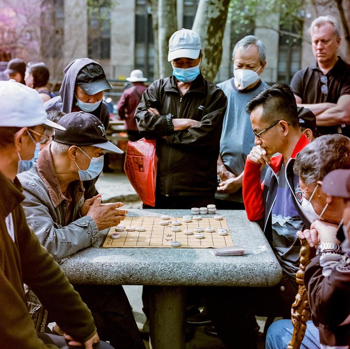 A group of men wearing face masks are gathered around a stone table playing a board game outdoors.