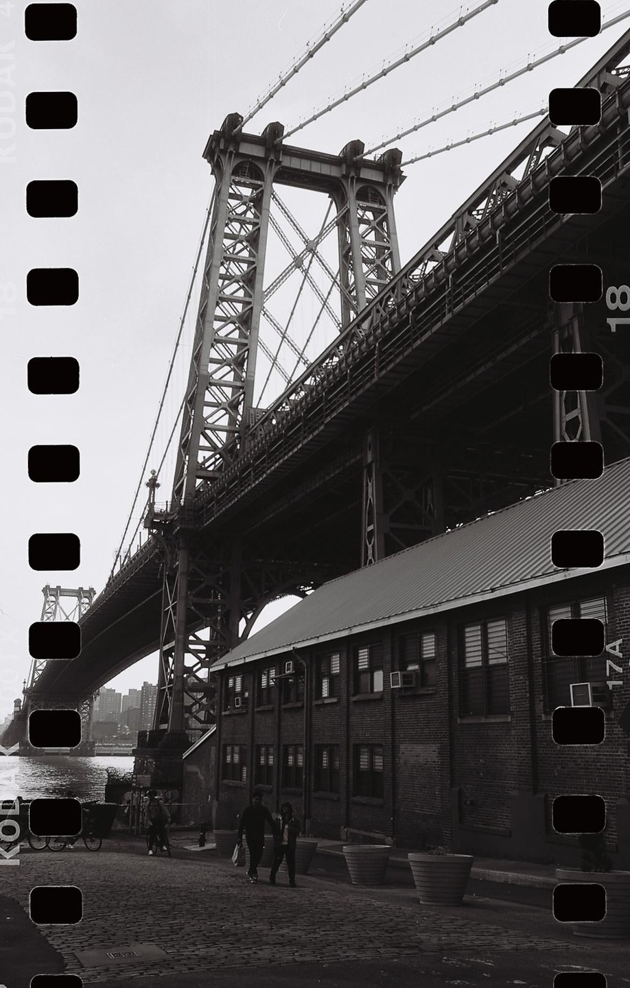 A large bridge spans over a brick building and a cobblestone street where two people walk.