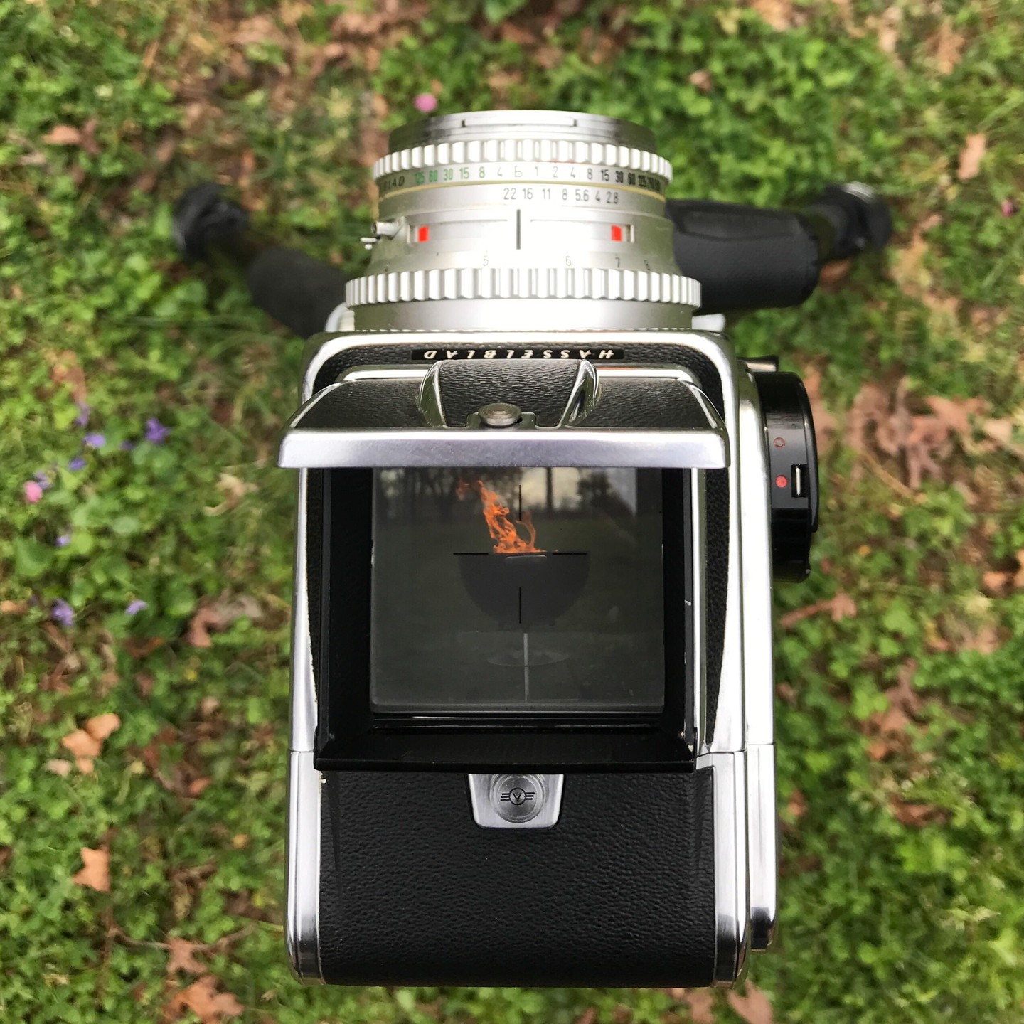 A camera with its viewfinder open is placed on green grass, showing a reflection of a fire pit with flames.