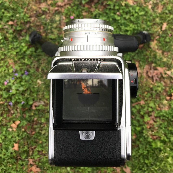 A camera with its viewfinder open is placed on green grass, showing a reflection of a fire pit with flames.