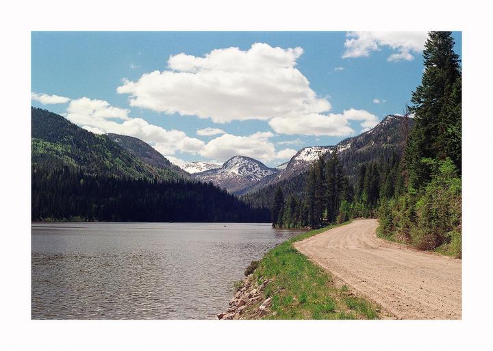 A dirt road curves along a lake with mountains and pine trees under a cloudy sky.