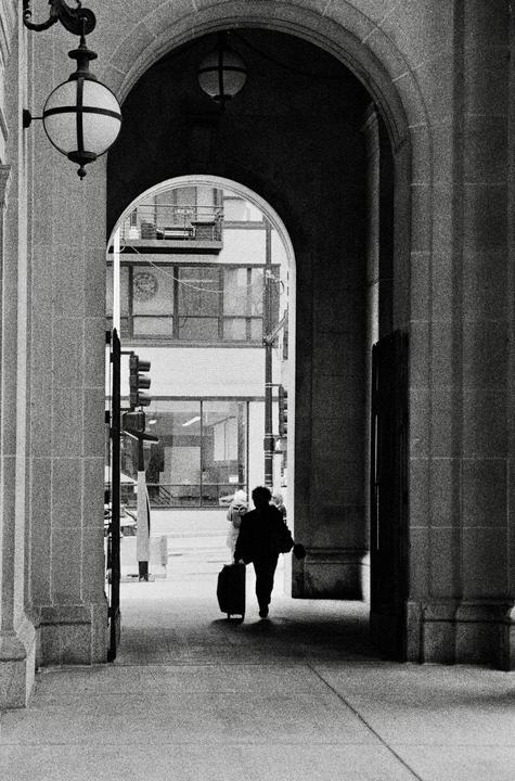 A person pulls a suitcase through an arched walkway, with a street and buildings visible in the background.