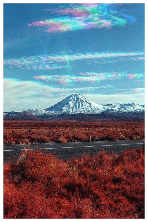 A snow-capped mountain rises above a road and red foliage under a blue sky with colorful clouds.