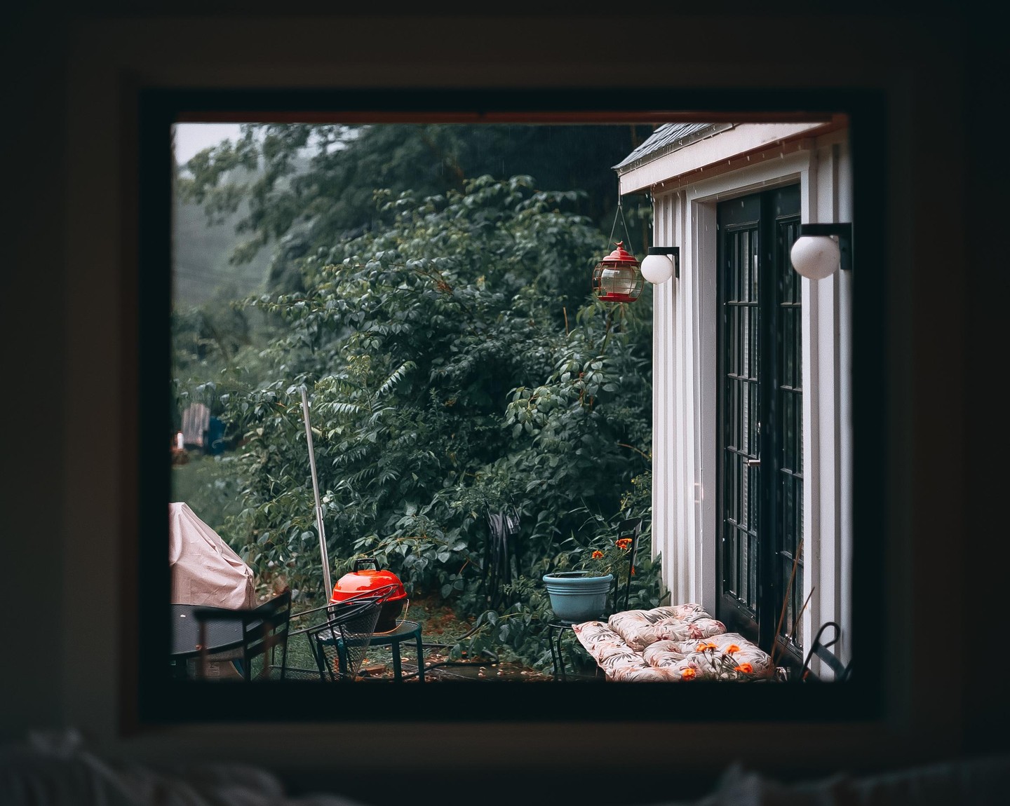 A red grill and a white building with a black door are visible through a window, surrounded by lush green foliage.