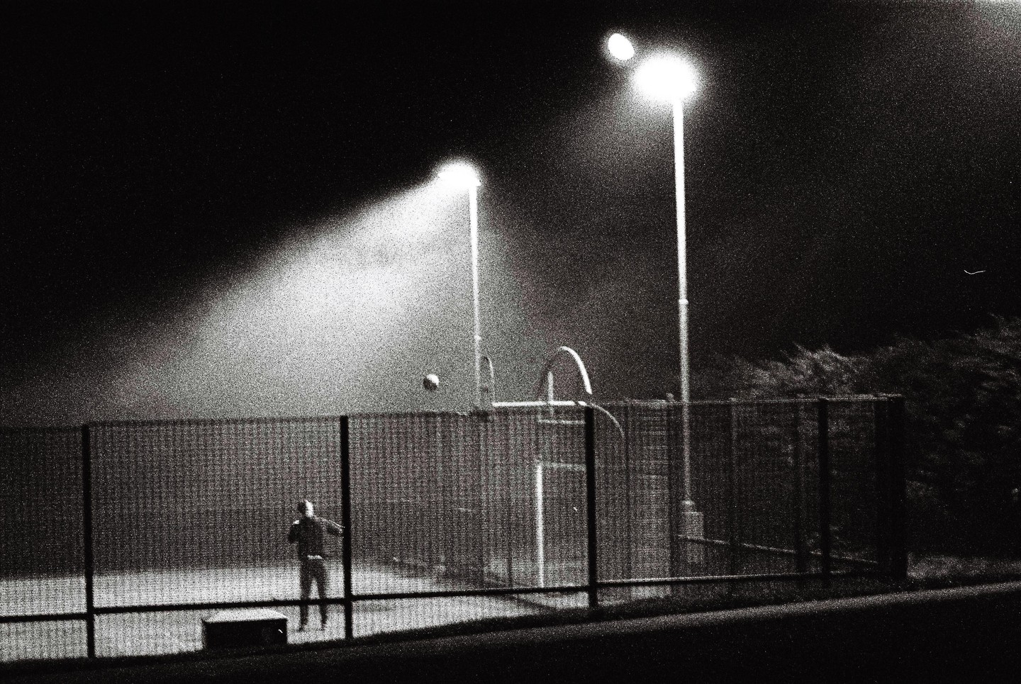 A person stands on a basketball court at night, illuminated by two bright lights, with a basketball in mid-air.