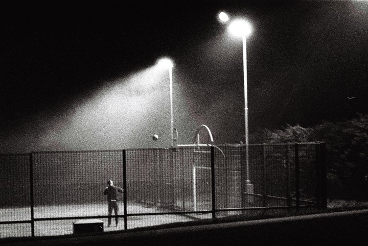 A person stands on a basketball court at night, illuminated by two bright lights, with a basketball in mid-air.
