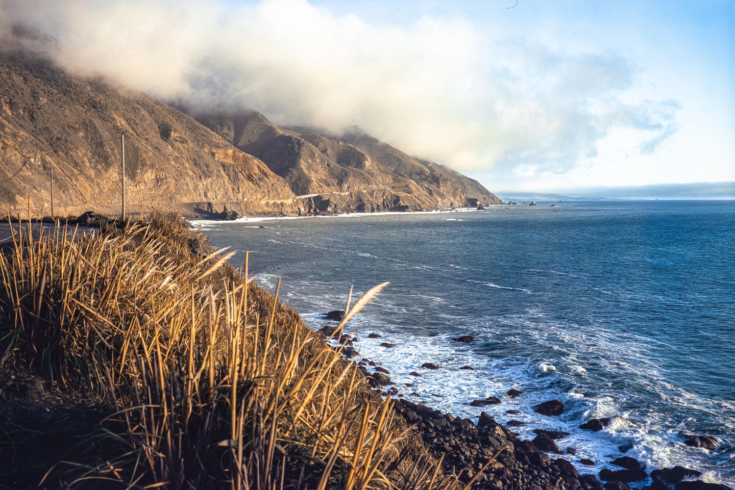 A coastal highway winds along a mountain range partially obscured by clouds, overlooking the Pacific Ocean with waves crashing on the rocky shore.