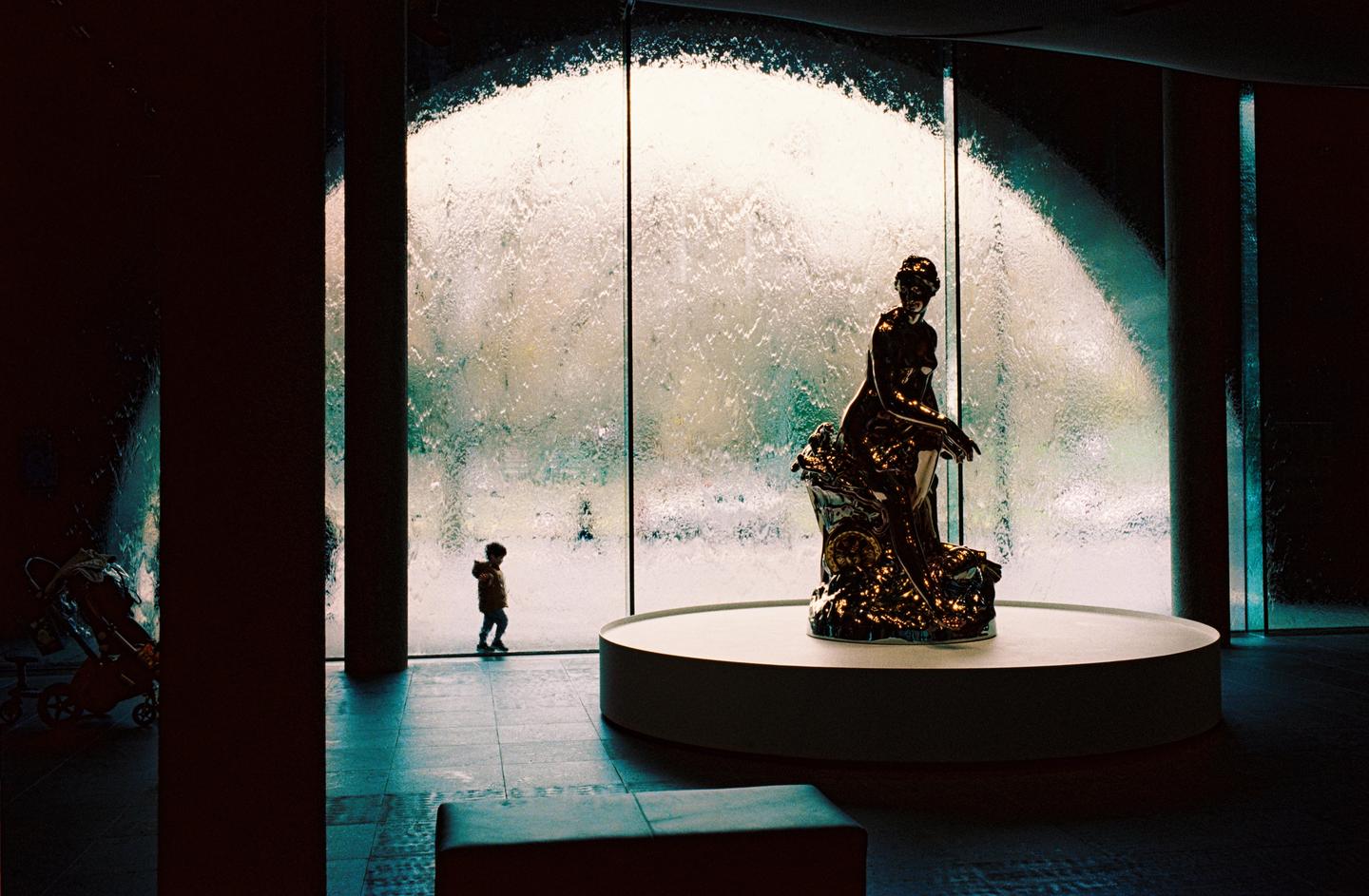 A child stands in front of a large window with water flowing down it, next to a statue of a woman.