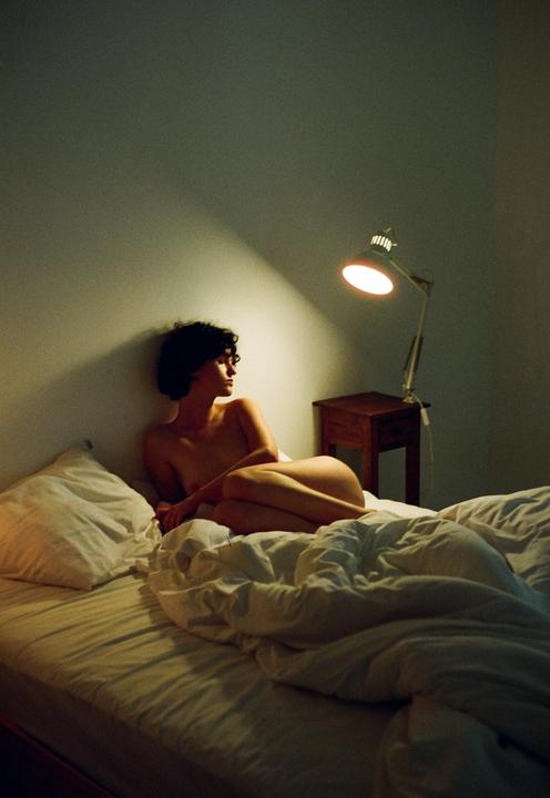 A woman with short, curly hair is lying on a bed, illuminated by a lamp on a bedside table.
