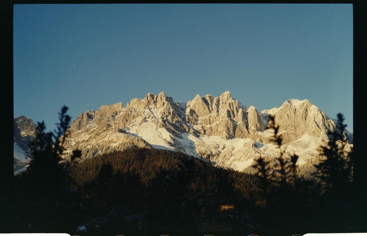 A snow-covered mountain range rises above a dark forest under a clear blue sky.