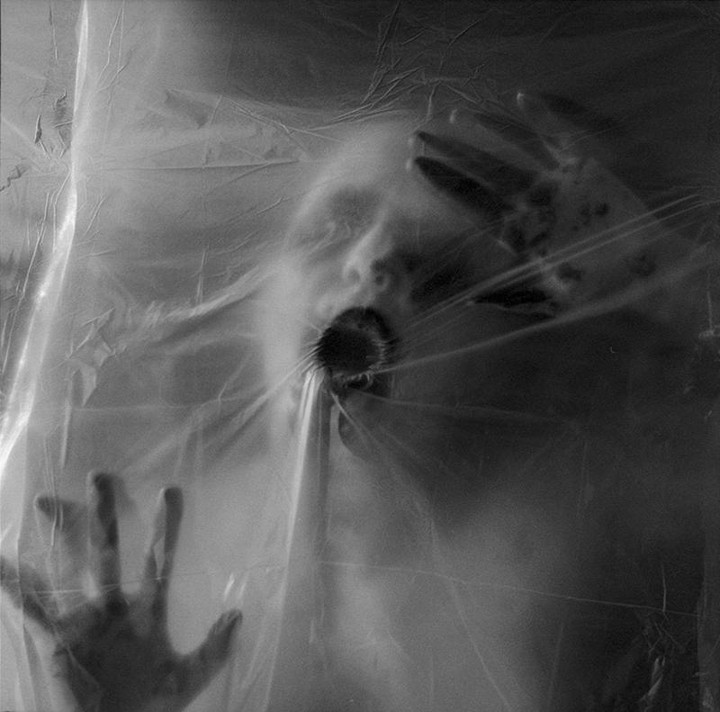 A person's face and hands are pressed against a translucent plastic sheet, with their mouth open.