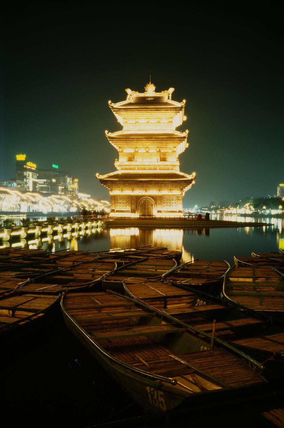 A brightly lit pagoda stands on the edge of a body of water with many small boats in the foreground at night.