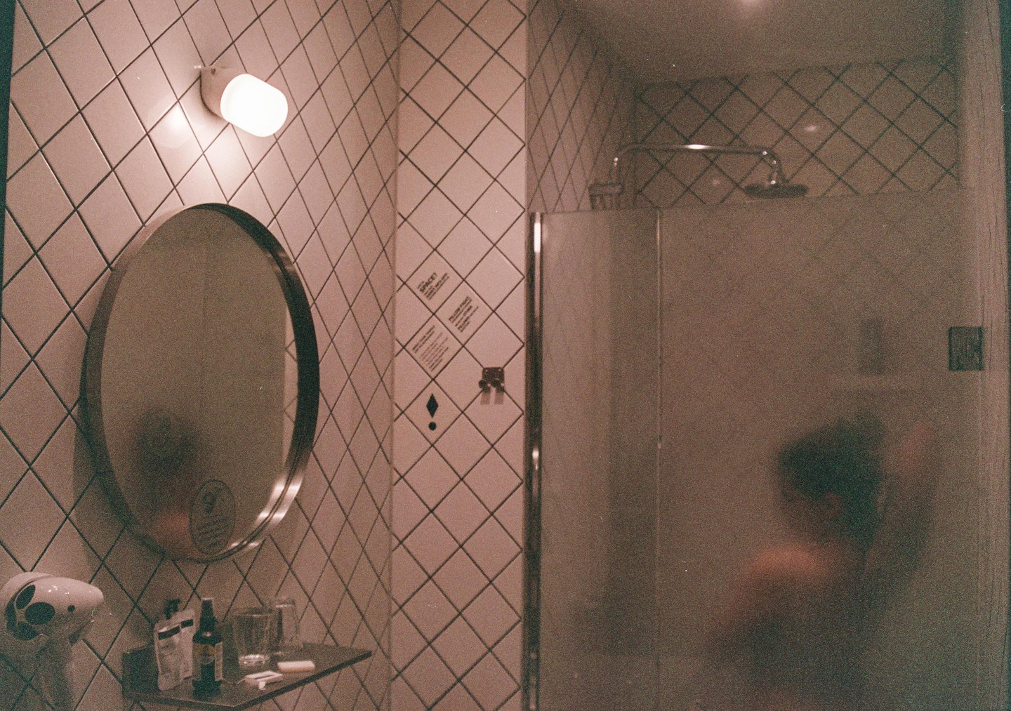 A person is showering behind a frosted glass door next to a mirror and tiled wall.