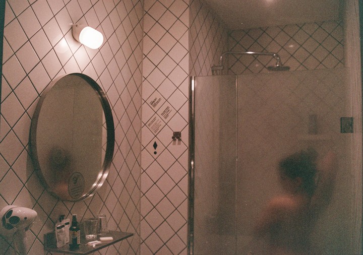A person is showering behind a frosted glass door next to a mirror and tiled wall.