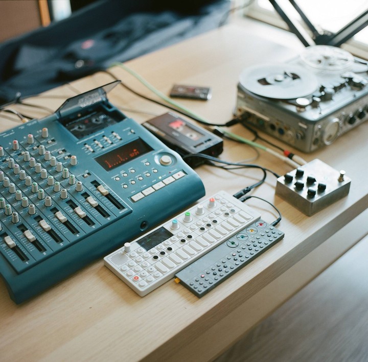 A collection of audio equipment, including a mixing board, a reel-to-reel player, and synthesizers, is arranged on a wooden table.