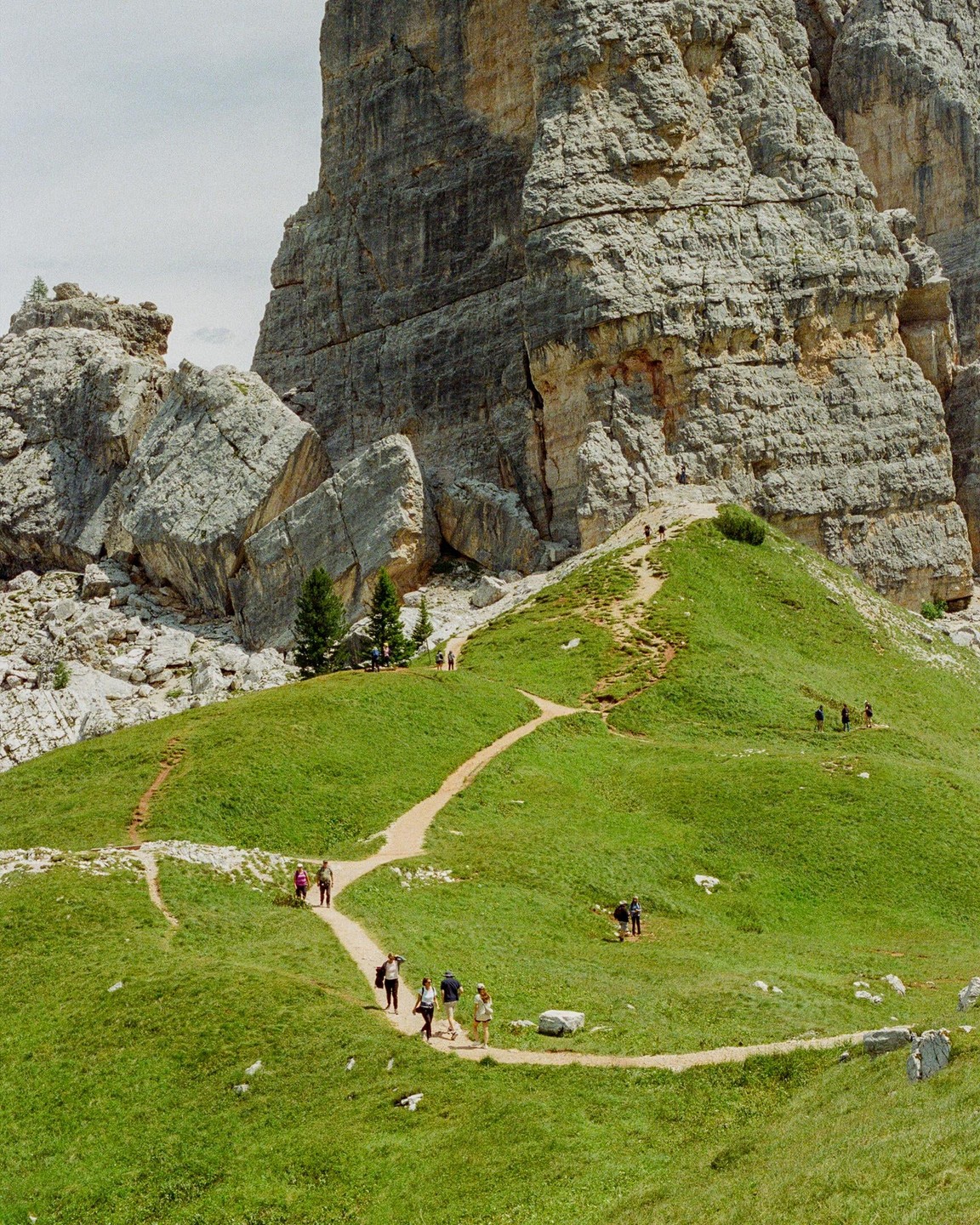 A group of hikers walks along a winding dirt path on a grassy hill with a large rock formation in the background.