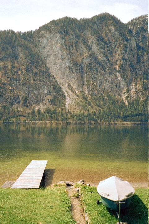 A small boat on a trailer sits on a grassy bank next to a lake with a wooden dock and a large mountain in the background.