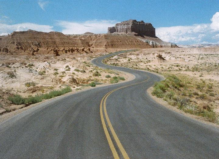 A winding road with double yellow lines leads through a desert landscape towards a large mesa under a blue sky with clouds.