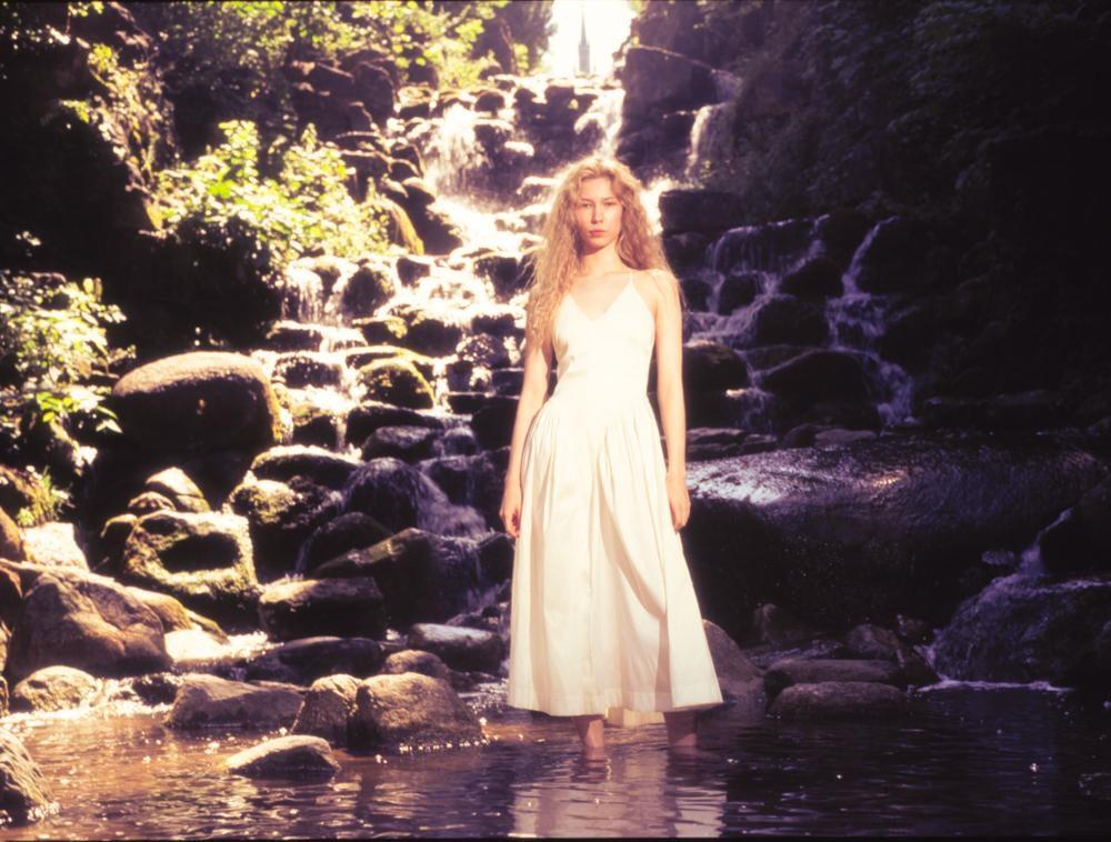 A woman in a white dress stands in a shallow stream with a waterfall and rocks behind her.