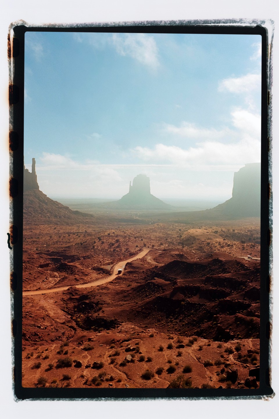 A winding dirt road leads through a vast desert landscape with towering rock formations under a partly cloudy sky.