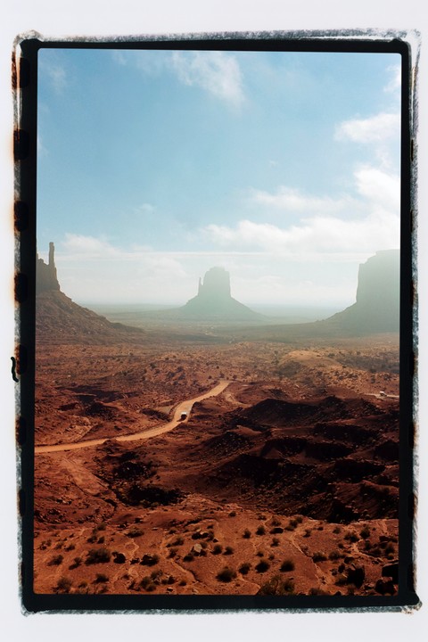 A winding dirt road leads through a vast desert landscape with towering rock formations under a partly cloudy sky.