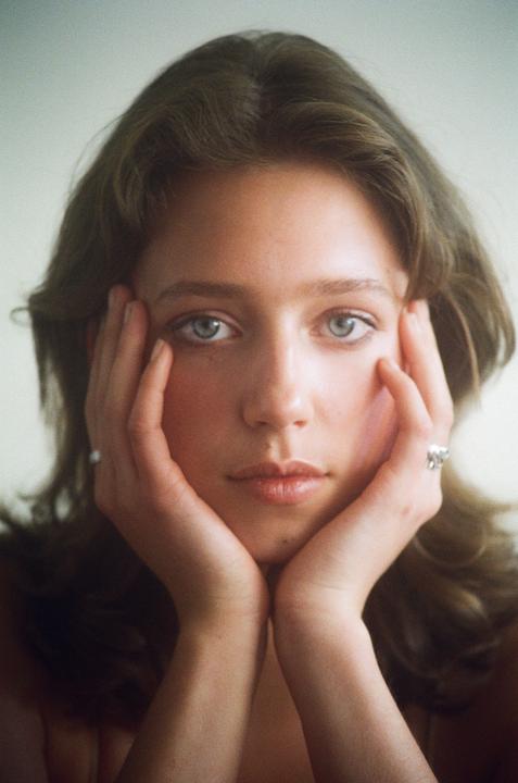 A woman with light eyes and brown hair rests her chin on her hands.