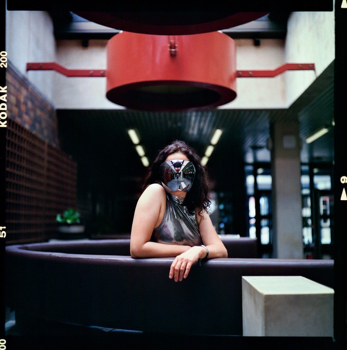 A woman with a reflective mask covering her face leans on a dark brown bench in a modern building.