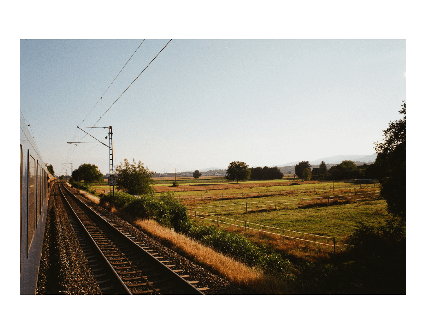 A train travels along a track next to a field with trees and hills in the distance.
